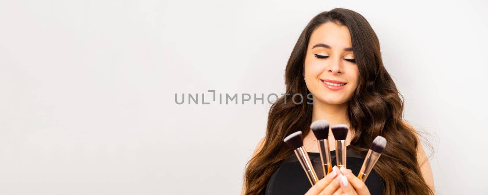 Young woman with makeup holding powder brush against grey background.