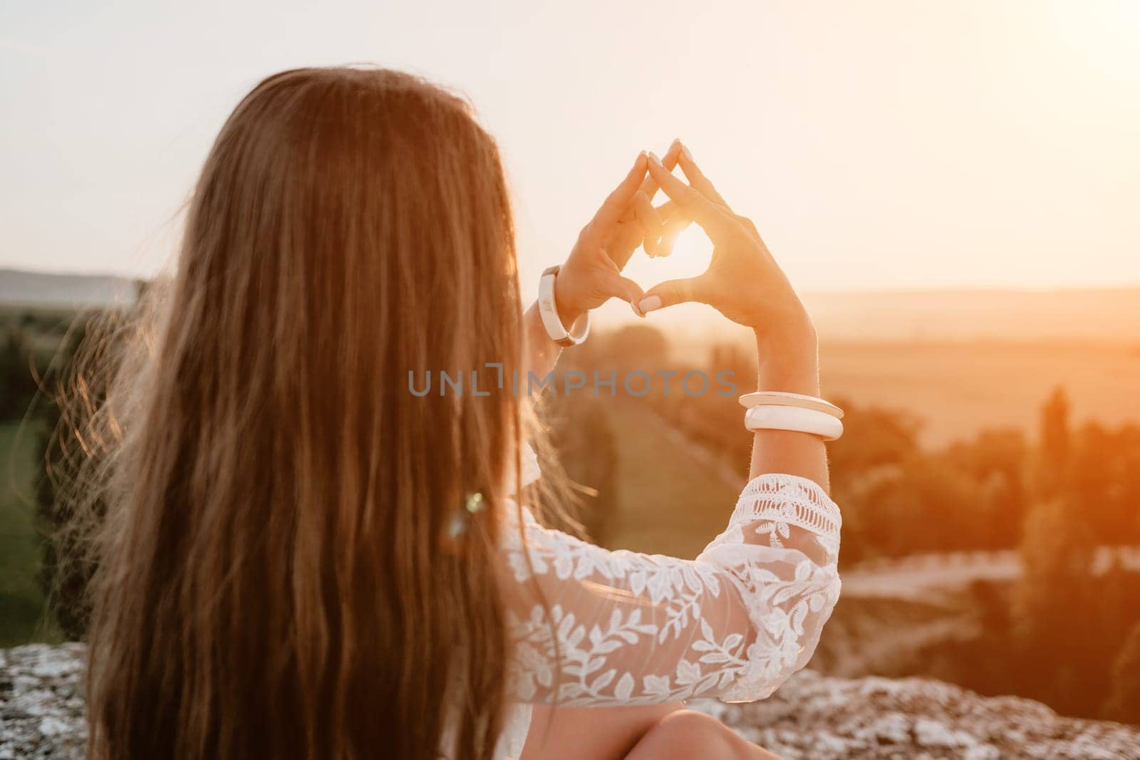 Happy woman in white boho dress making heart sign with hands on sunset in mountains. Romantic woman with long hair standing with her back on the sunset in nature in summer with open hands. by panophotograph