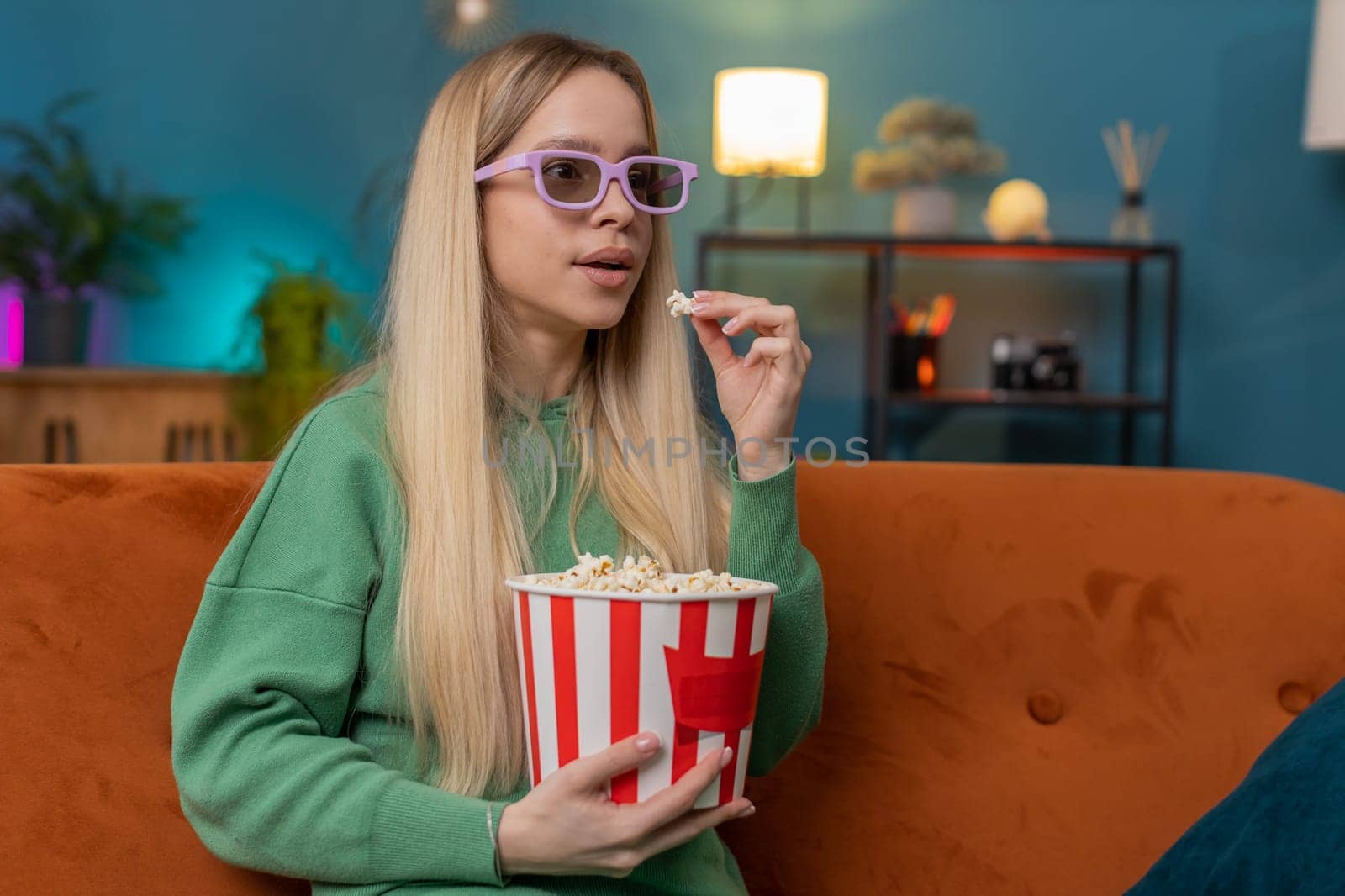 Excited woman in 3D glasses sits on couch eating popcorn snacks and watching interesting TV serial sport game, film online social media movie content at home apartment. Girl enjoying entertainment