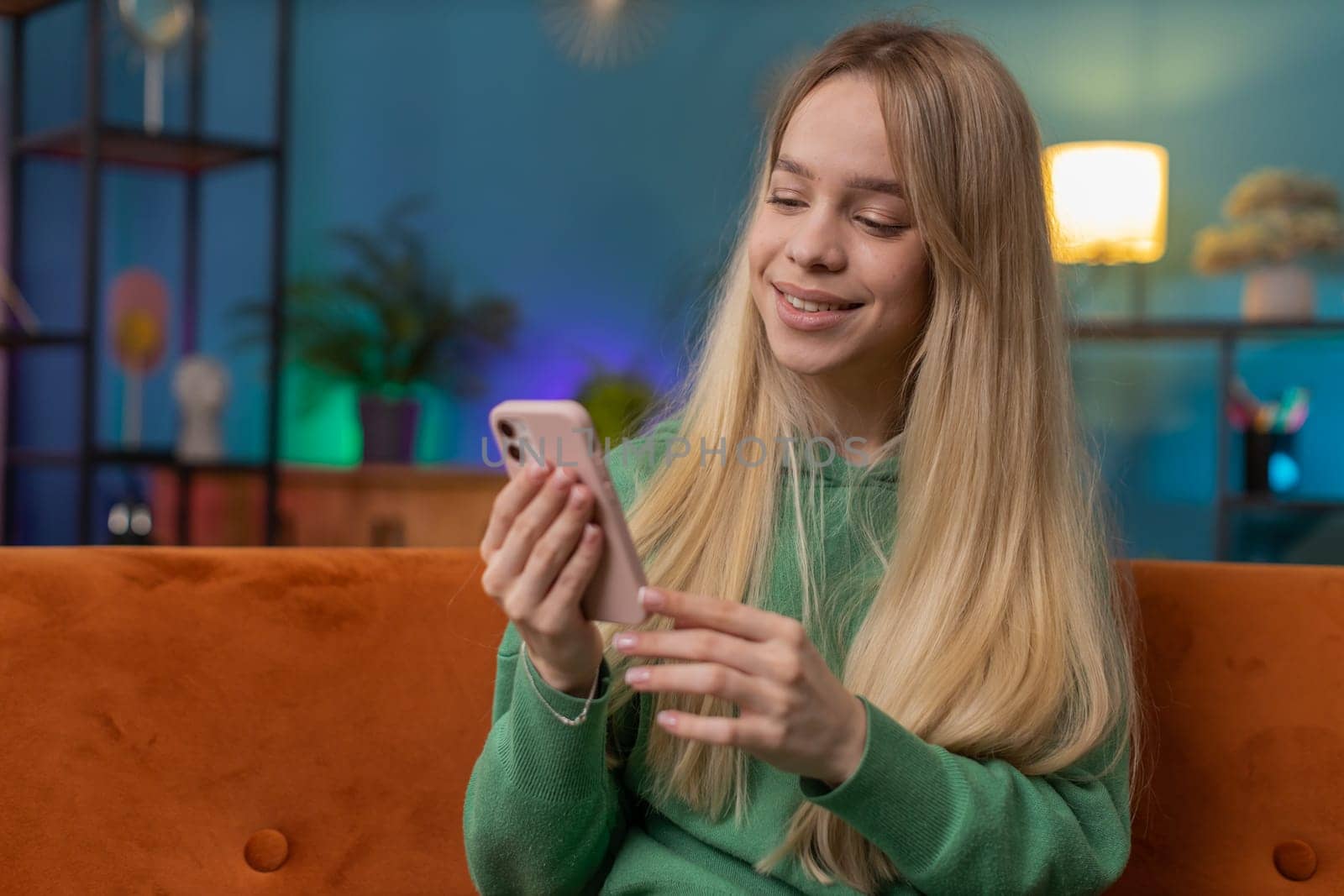 Caucasian woman sitting on couch uses mobile phone smiles at home living room apartment. Young girl texting share messages content on smartphone social media applications online, watching relax movie