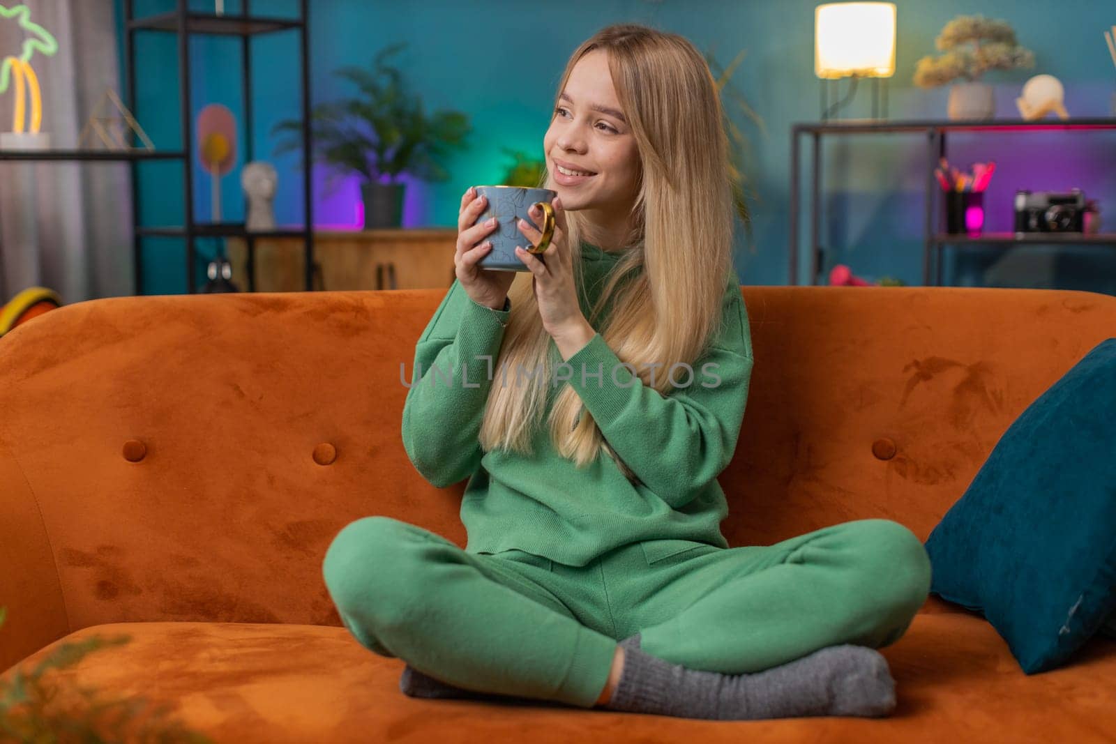 Portrait of smiling woman drinking a cup of warm coffee or herbal tea in the morning. Caucasian teen girl enjoying comfortable relaxing taking a break alone at home apartment living room on sofa