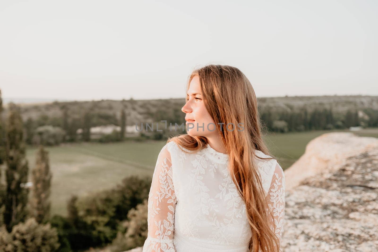 Romantic beautiful bride in white dress posing with sea and mountains in background. Stylish bride standing back on beautiful landscape of sea and mountains on sunset