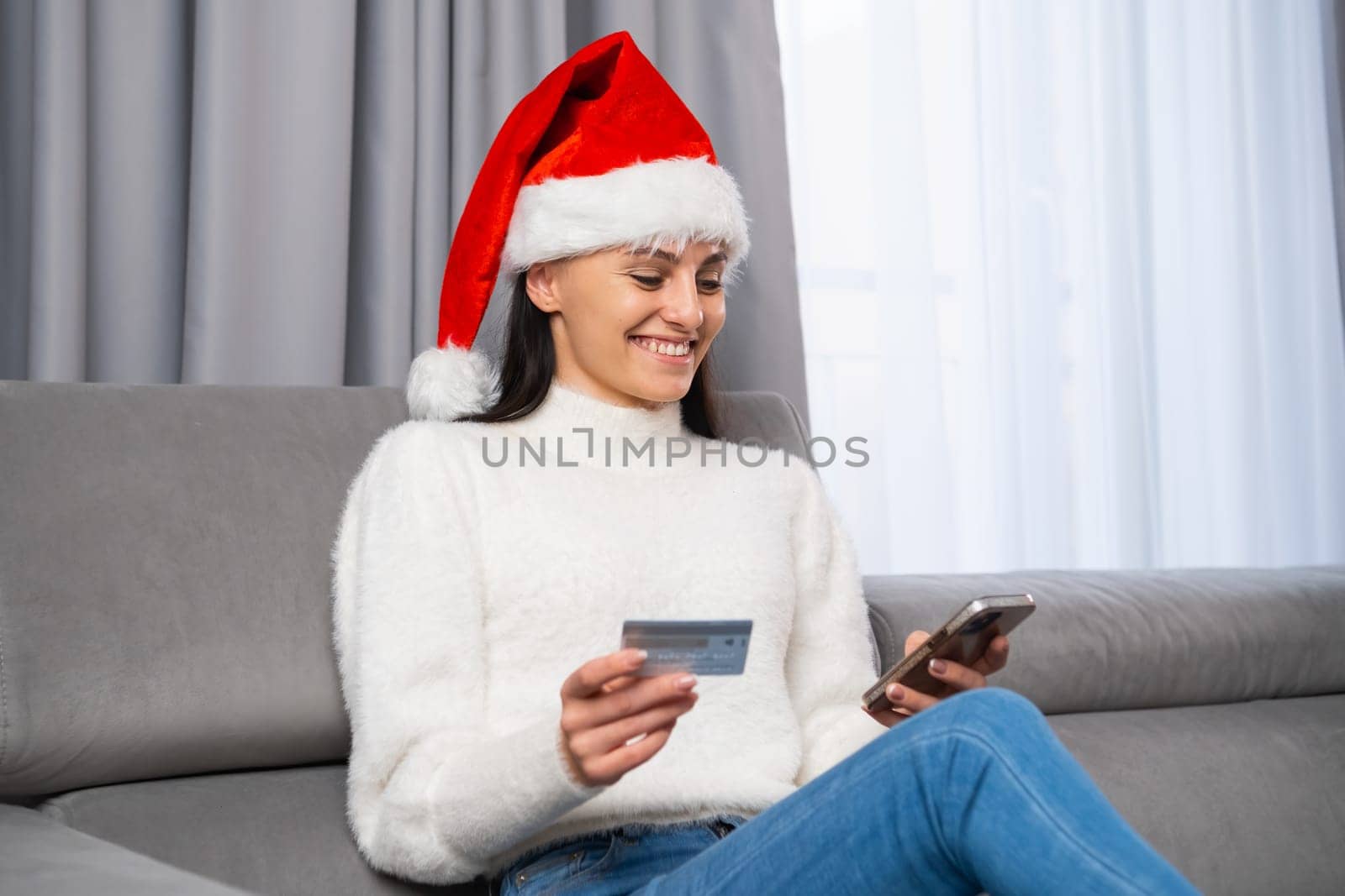 A woman in a red Santa Claus hat purchases Christmas gifts via the Internet using a mobile phone and credit card sitting in the living room.
