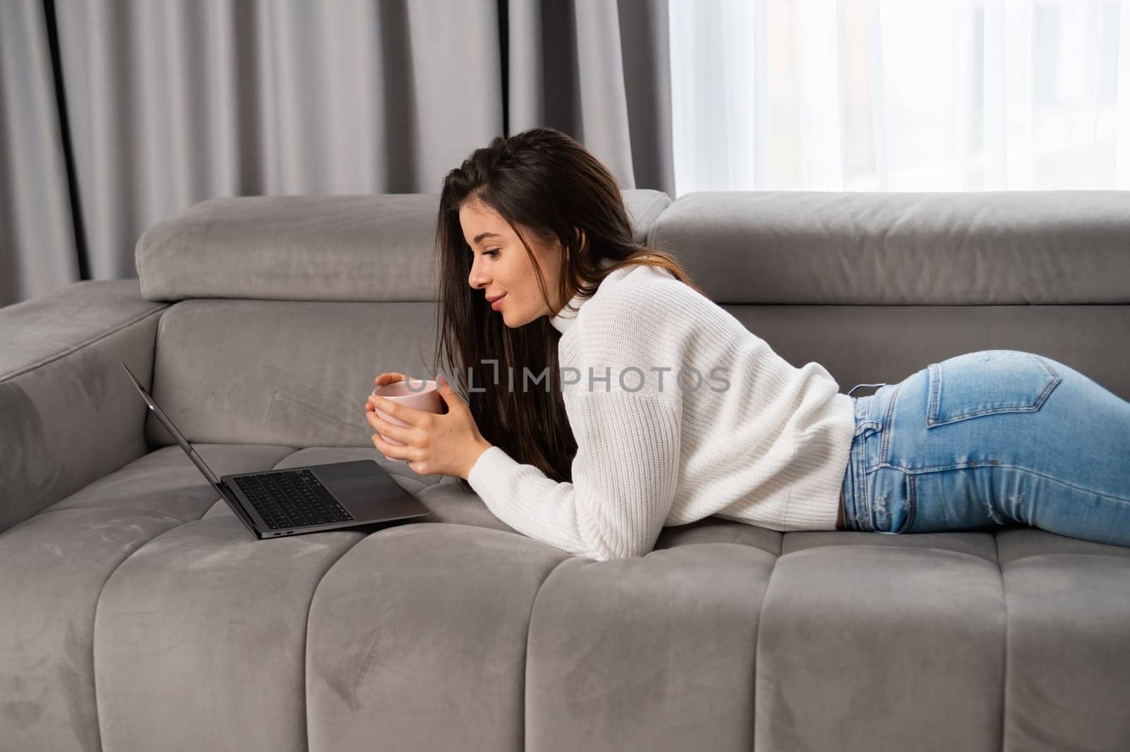 Young woman lying on couch with cup of tea in the living room and working online on the laptop.