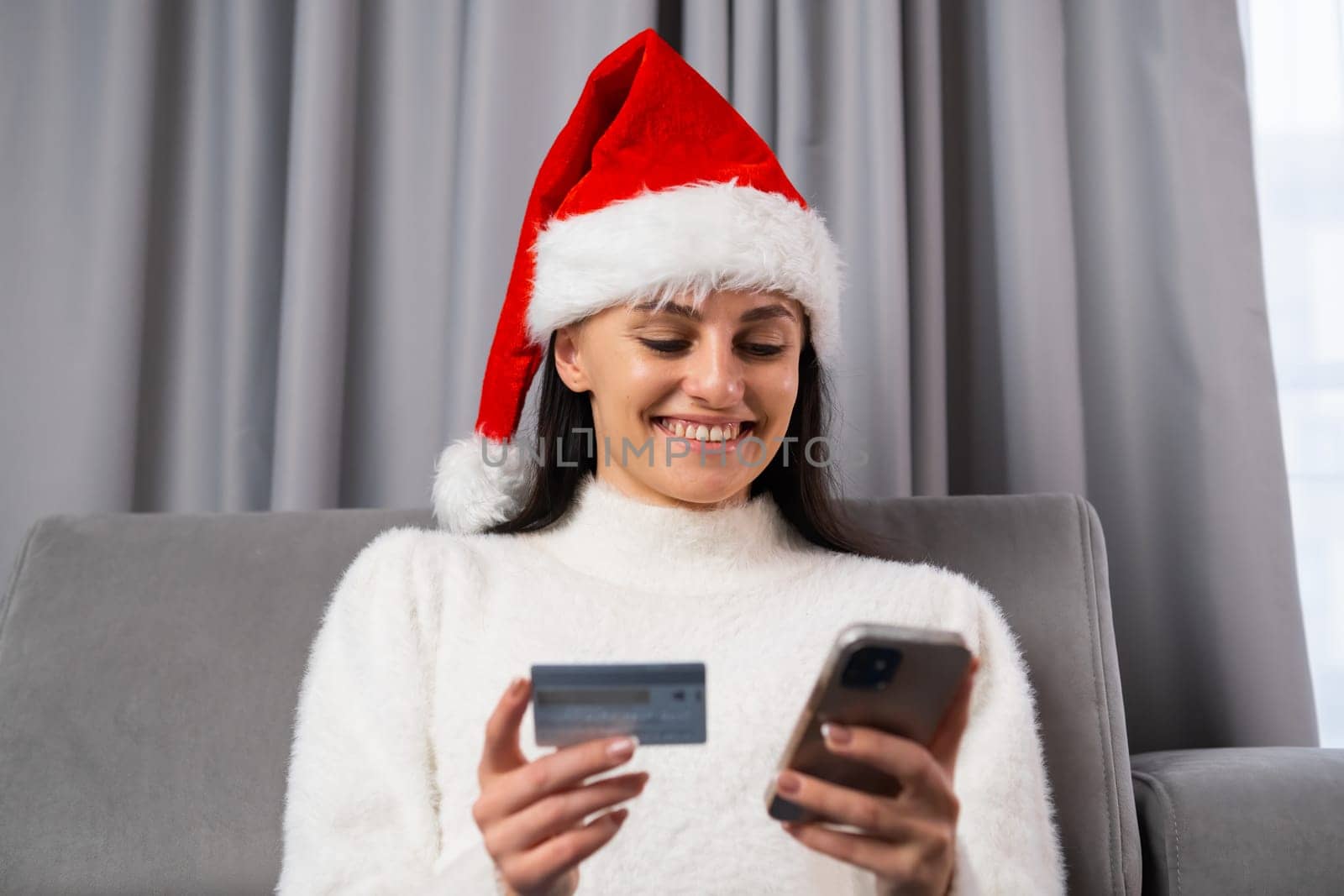 A young woman in Santa Claus hat orders gifts online. Spending money at home in a modern grey interior.