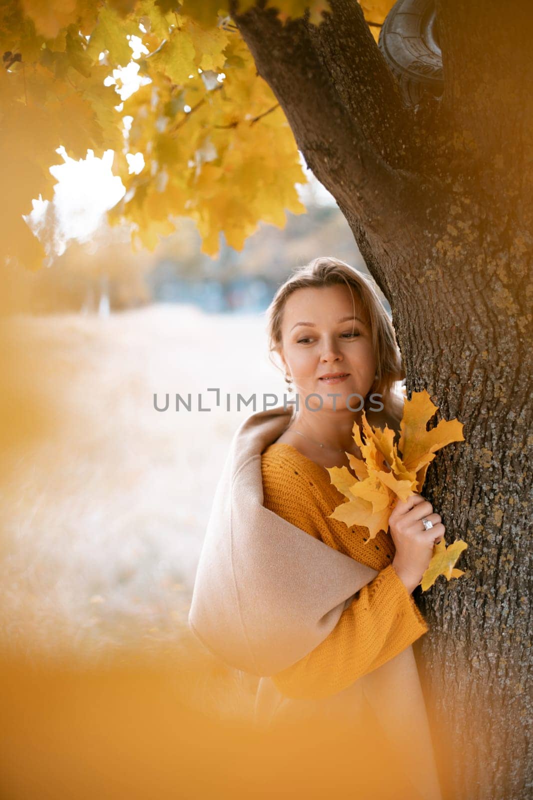 The blonde stands near the autumn tree. Thoughtful woman looks ahead, dressed in a yellow dress. Autumn content.