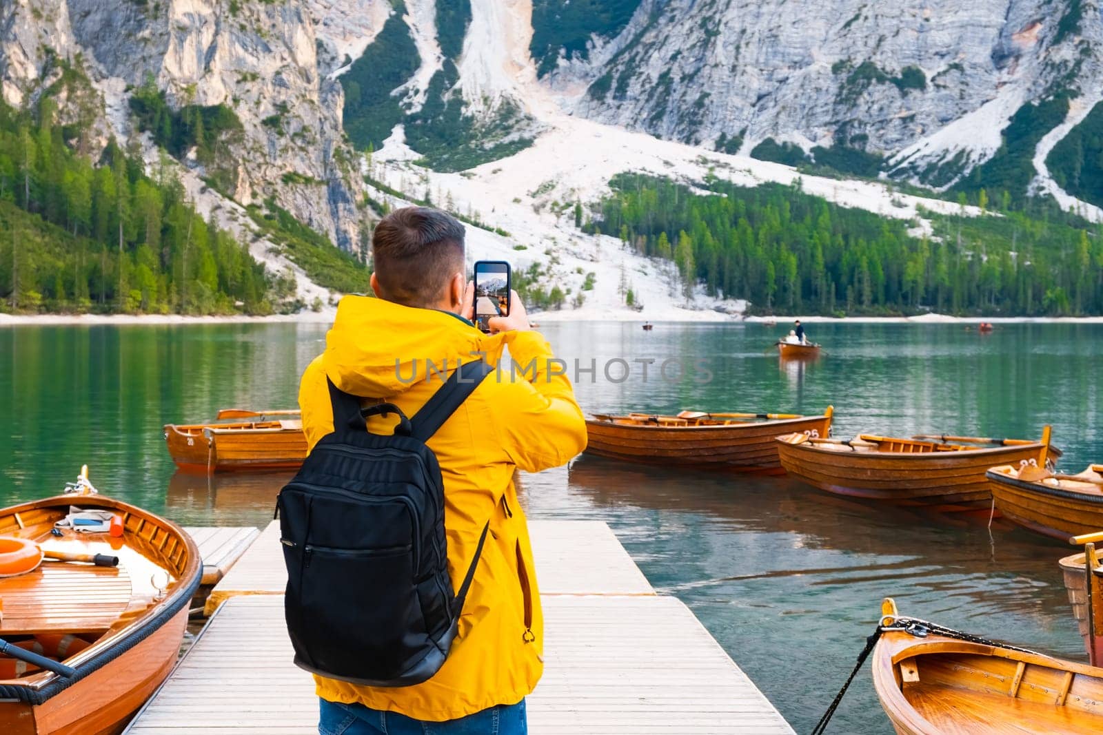 Tourist taking photo of wooden boats and Dolomites on the lake Braies. Man wearing yellow jacket and holding a black backpack visiting Dolomites Alps, Italy.