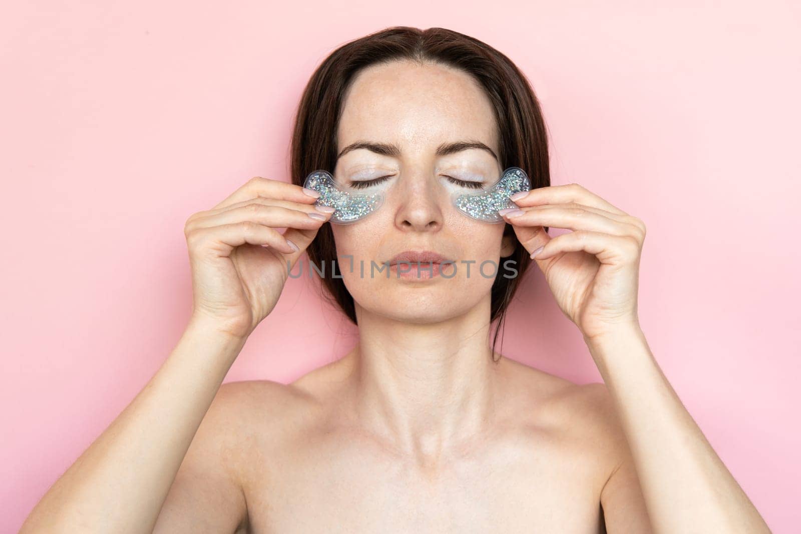 Pleased brunet woman closed eyes undergoes beauty procedures apply hydrogel patches under eyes to remove puffiness and wrinkles isolated over pink background. Skin care beauty treatment at home. by Ri6ka