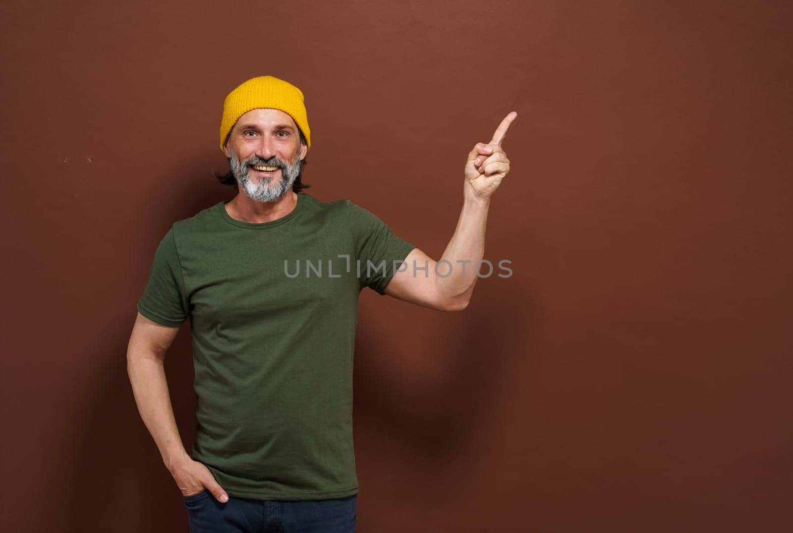 Smiling European man in a yellow hat points his hand to the side with copy space on a brown background. Product placing, advertising concept. by LipikStockMedia