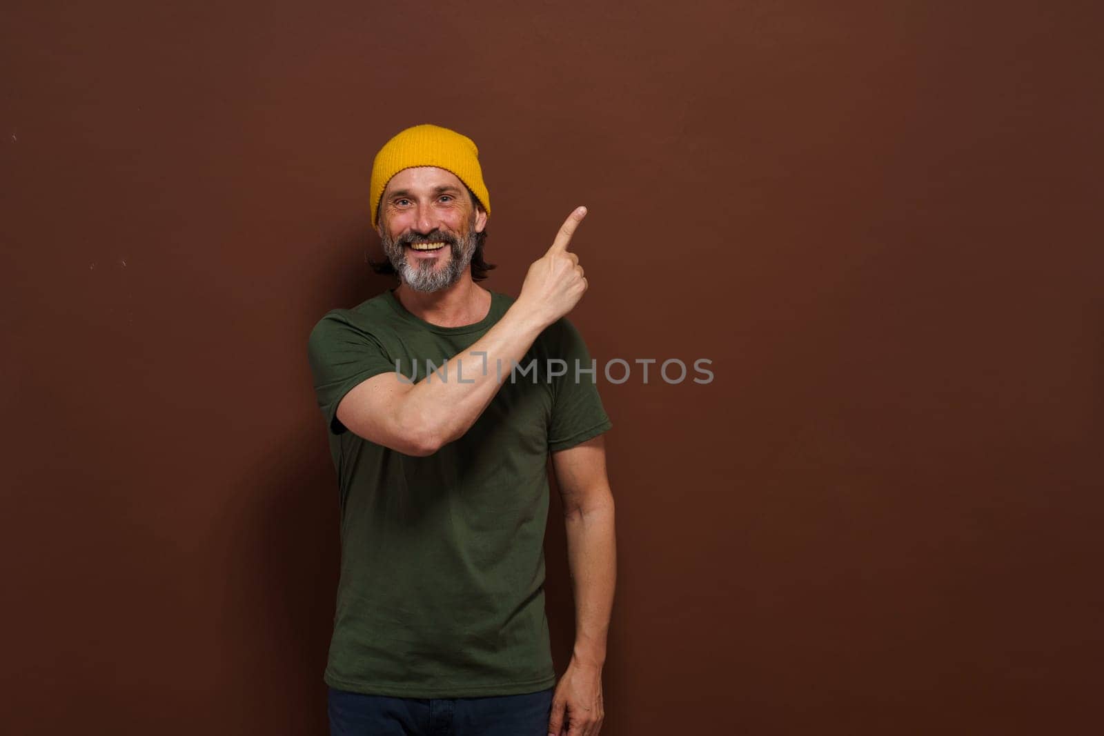 Excited smiling Caucasian man in a yellow hat points his hand to the side with copy space on a brown background. Product placing, advertising concept. High quality photo