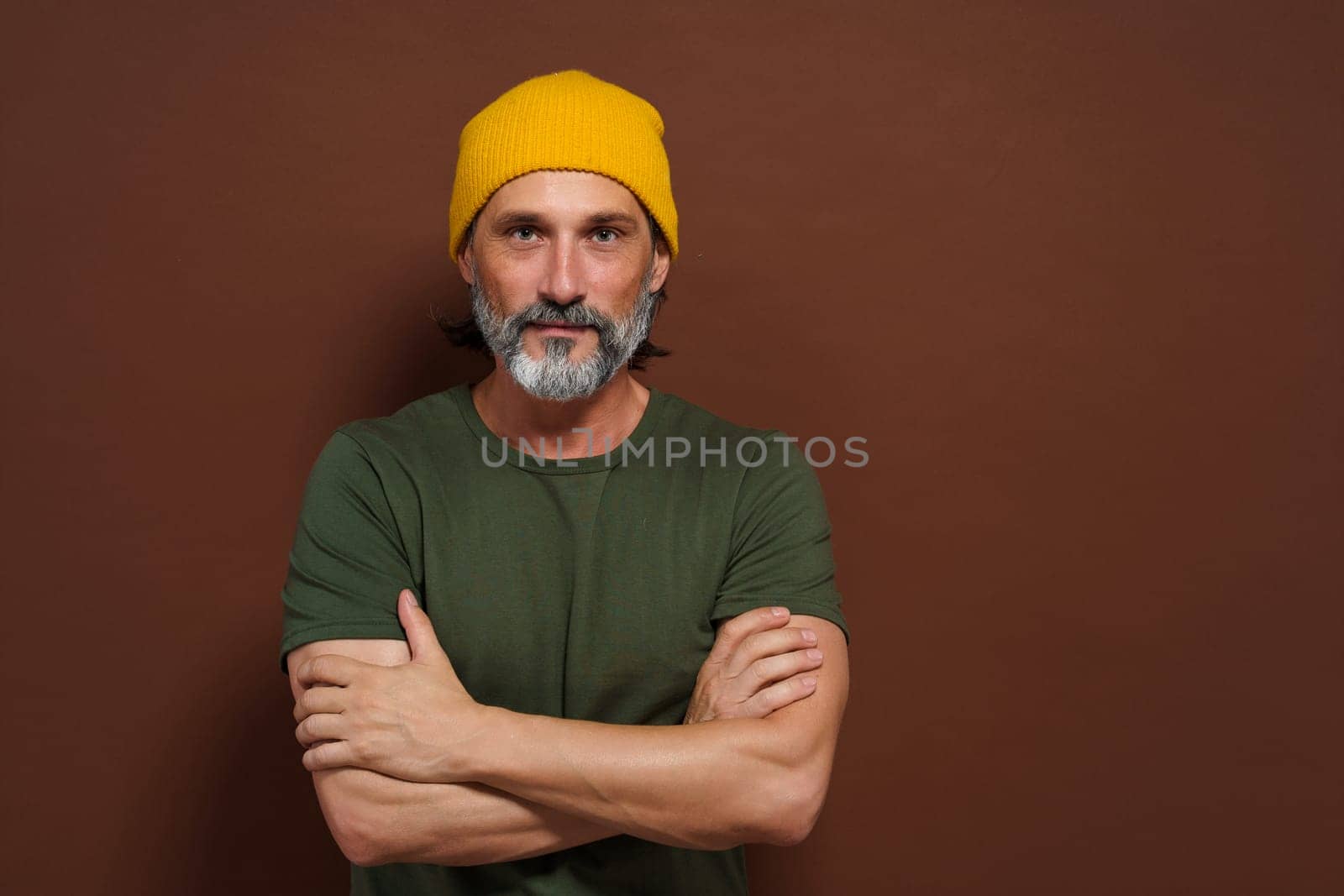 Mature, handsome man with silver beard wearing yellow hat against brown background. The image has copy space and can be used for product placement or advertising concepts. by LipikStockMedia