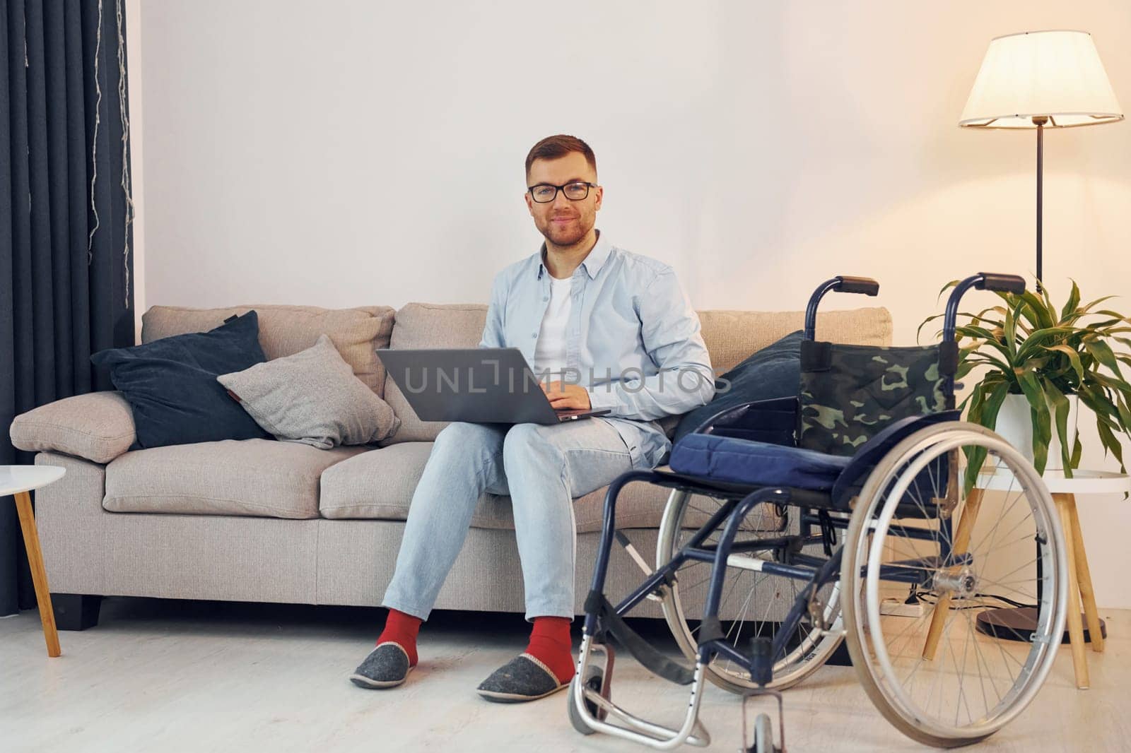 With laptop on the legs. Disabled man in wheelchair is at home.