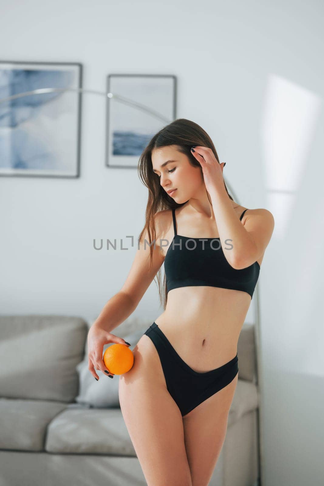 With orange in hand. Beautiful woman in underwear is posing indoors.