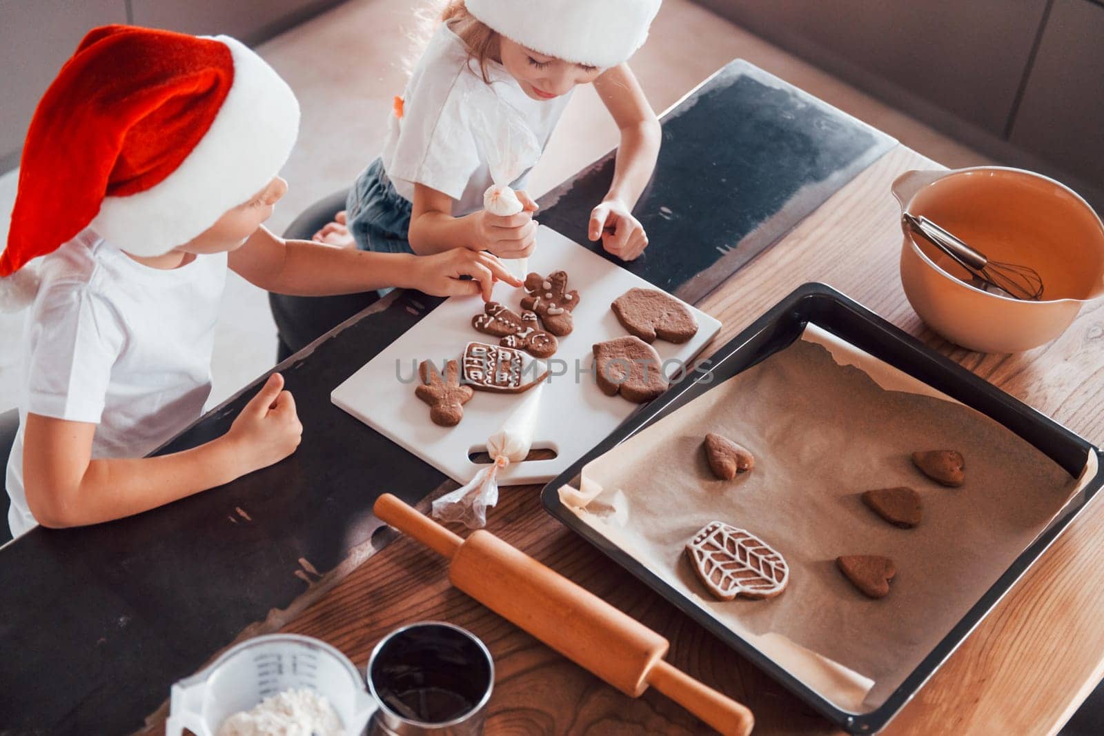In santa hats. Little boy and girl preparing Christmas cookies on the kitchen by Standret
