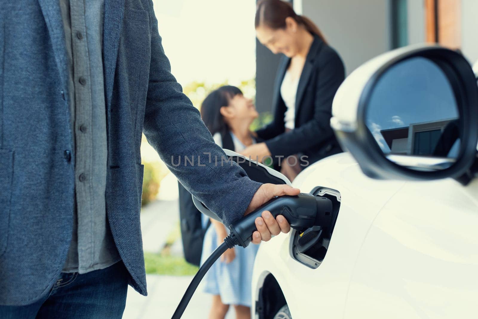 Closeup focus hand insert EV charger plug to electric vehicle at home charging station with blurred family in background. Progressive concept of green and clean energy to reduce CO2 emission by EV car