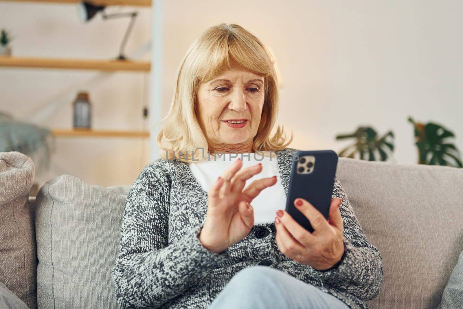 Sitting and holding phone. Senior woman with blonde hair is at home.