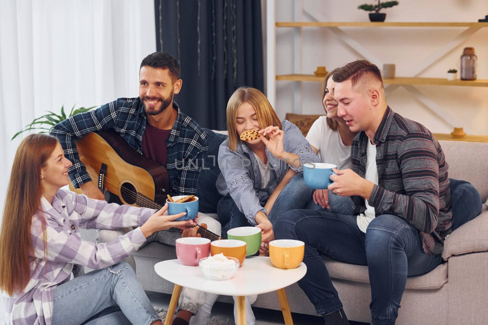 Man is playing acoustic guitar. Group of friends have party indoors together.