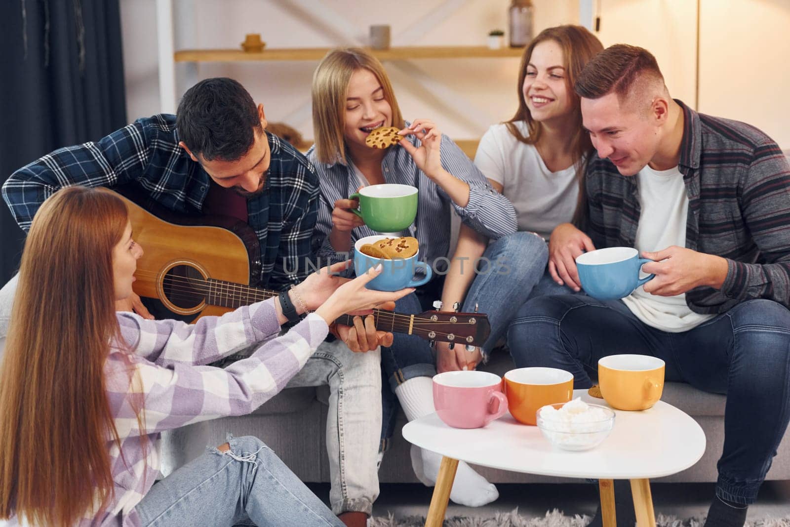 Man is playing acoustic guitar. Group of friends have party indoors together.