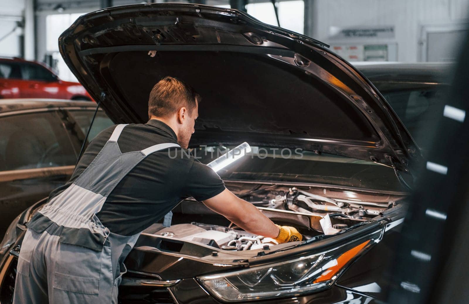 With lighting equipment. Man in uniform is working in the auto service.