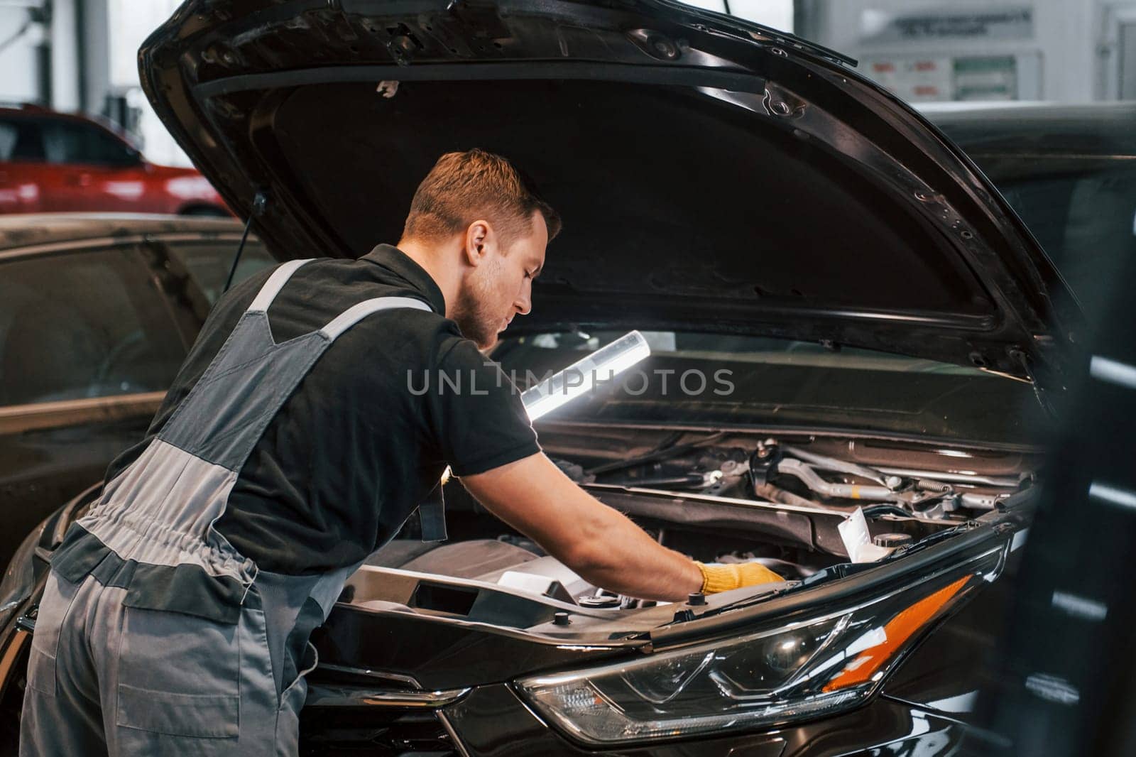 With lighting equipment. Man in uniform is working in the auto service.