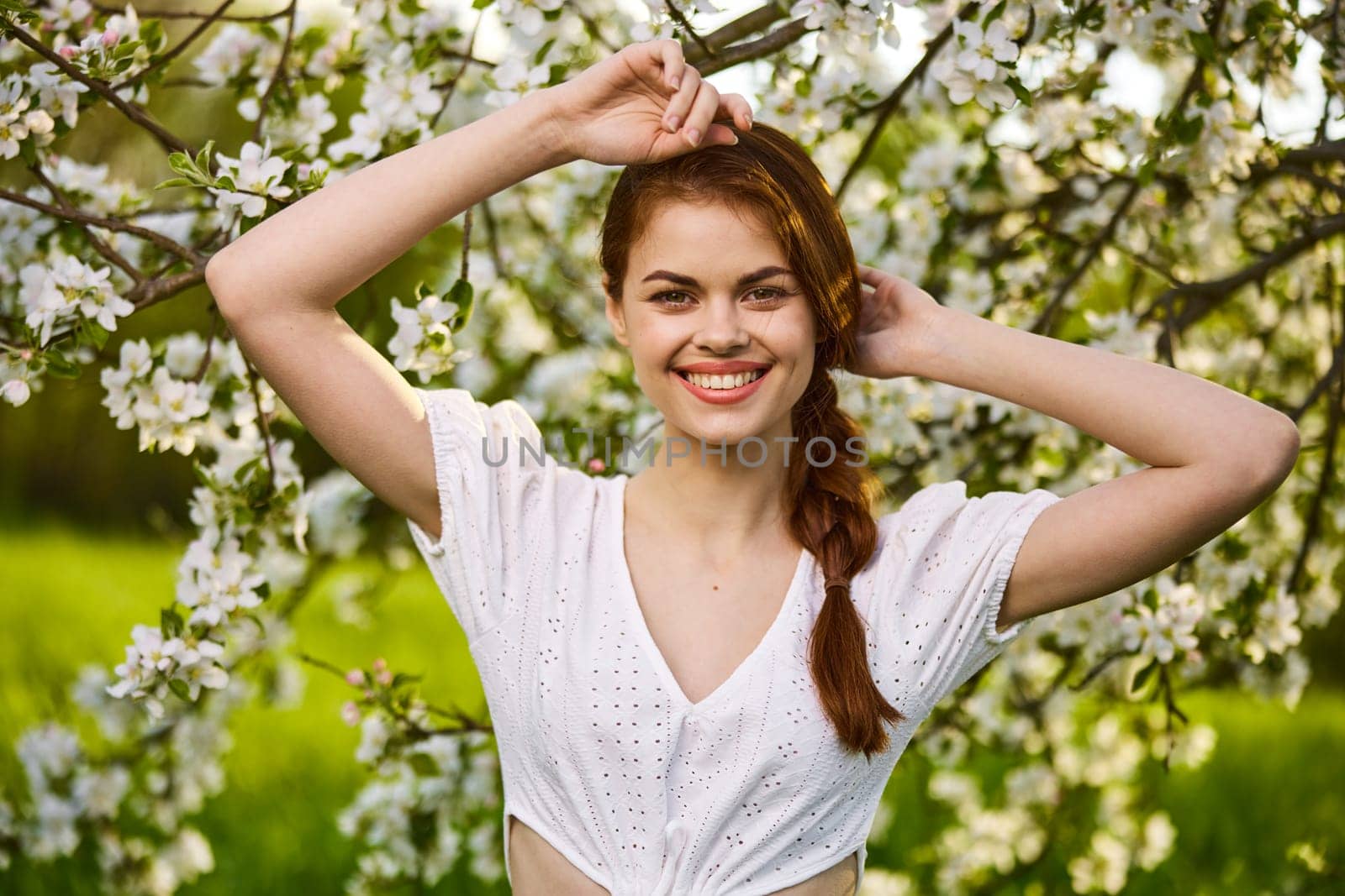 portrait of a joyful woman in a light dress against the background of a flowering tree, raising her hands to her head by Vichizh