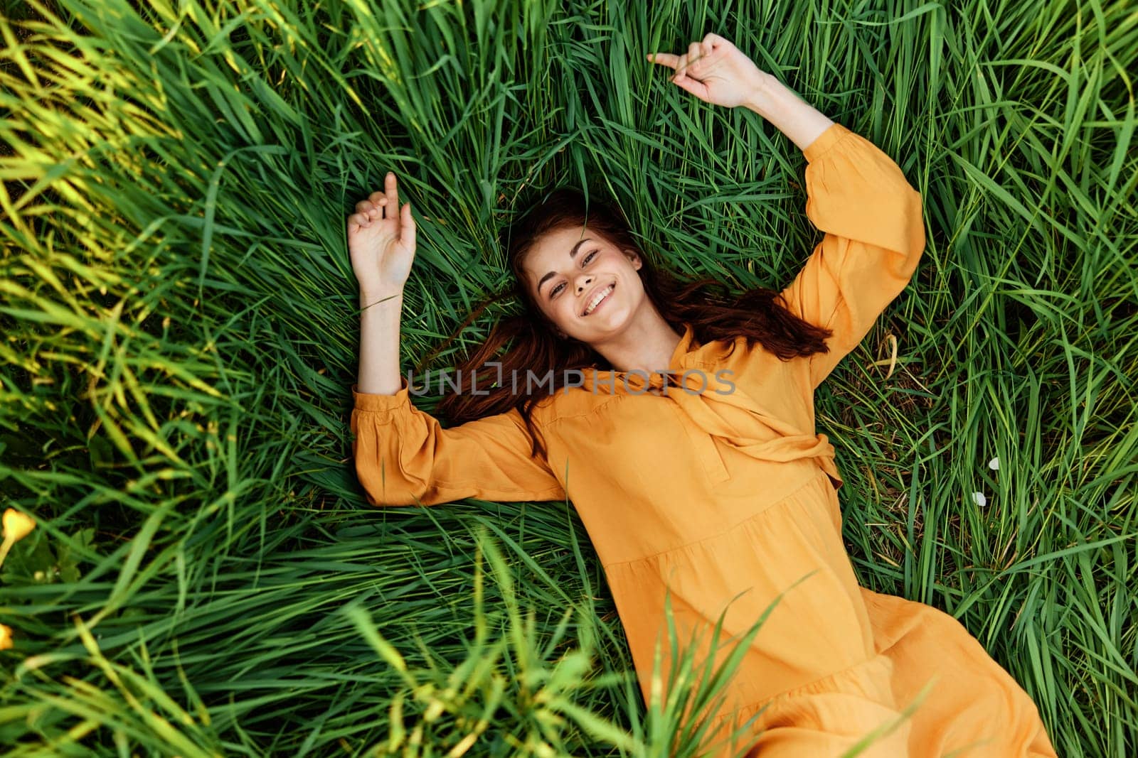 a joyful woman in a long orange dress lies in the tall green grass and looking at the camera smiles broadly. High quality photo