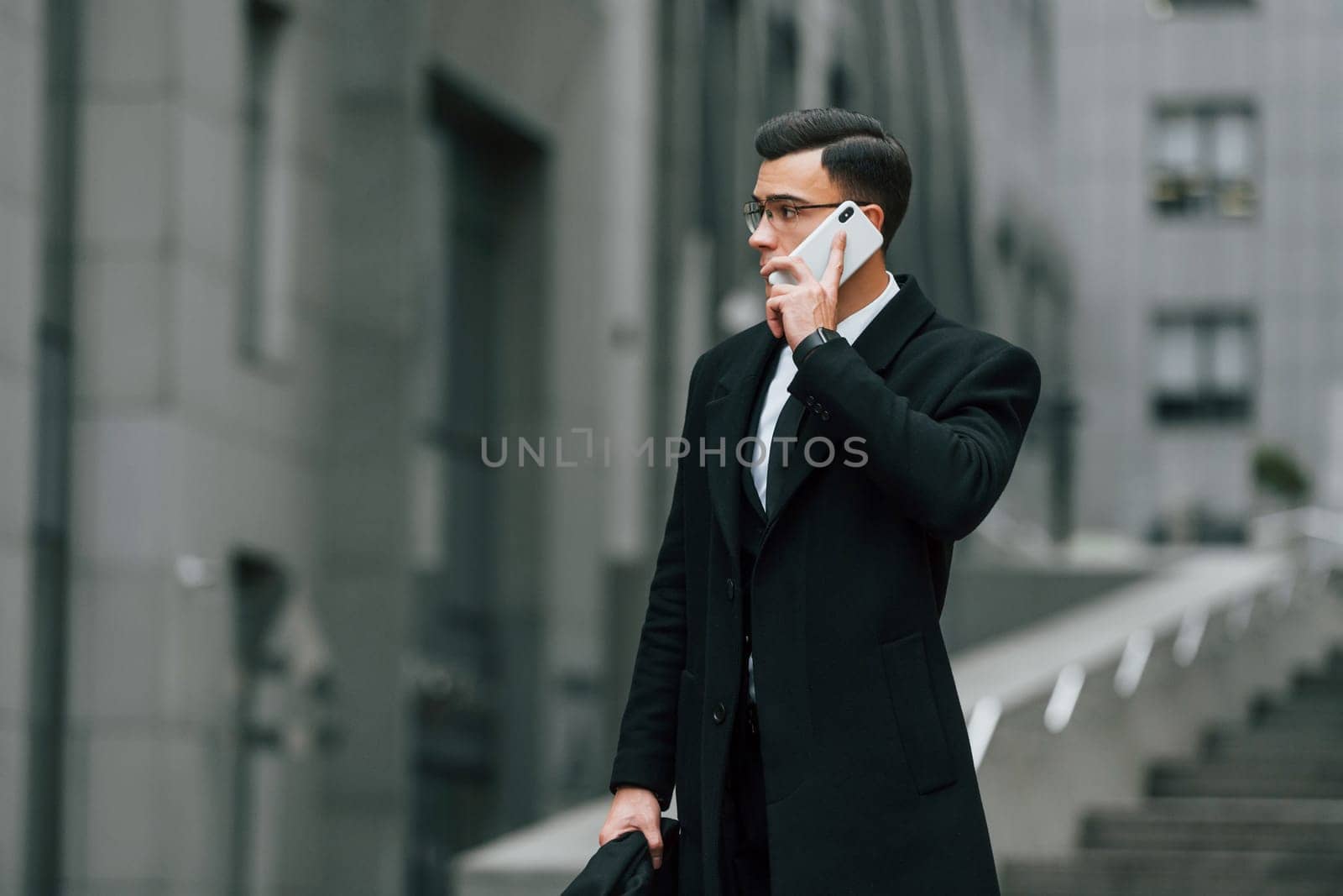 Talking by phone. Businessman in black suit and tie is outdoors in the city by Standret
