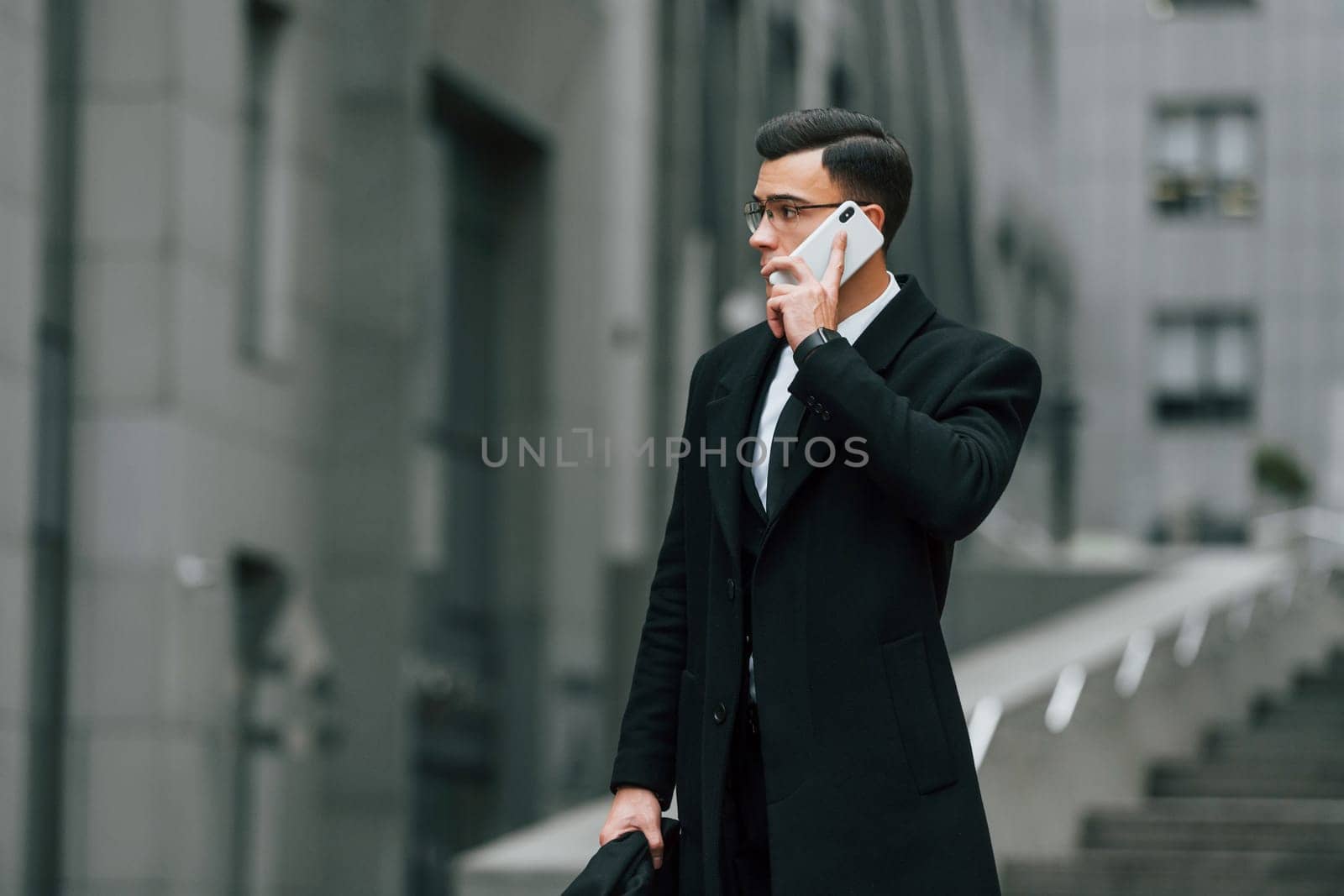 Talking by phone. Businessman in black suit and tie is outdoors in the city.