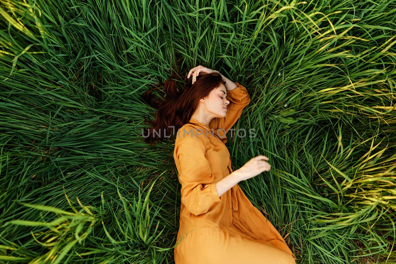 an elegant woman lies in the tall green grass in a long summer orange dress and holding her hair with her hands enjoys nature and sunny weather with her eyes closed by Vichizh