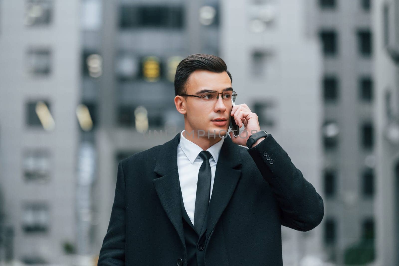 Talking by phone. Businessman in black suit and tie is outdoors in the city.