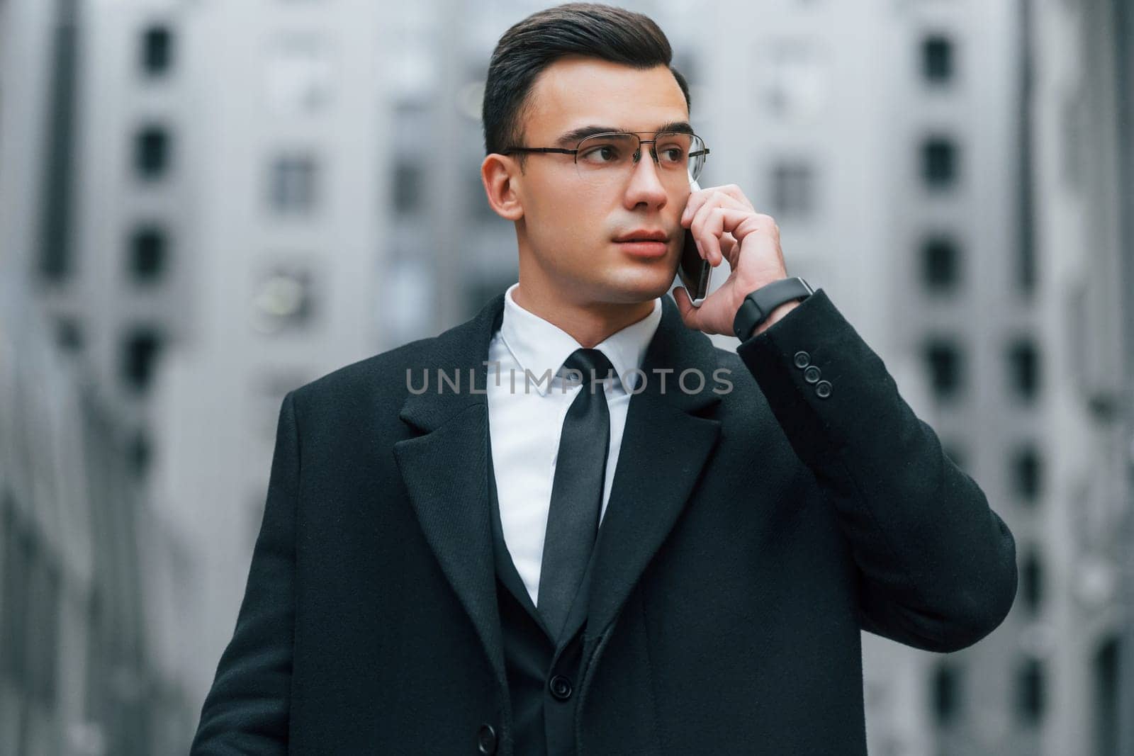Talking by phone. Businessman in black suit and tie is outdoors in the city.