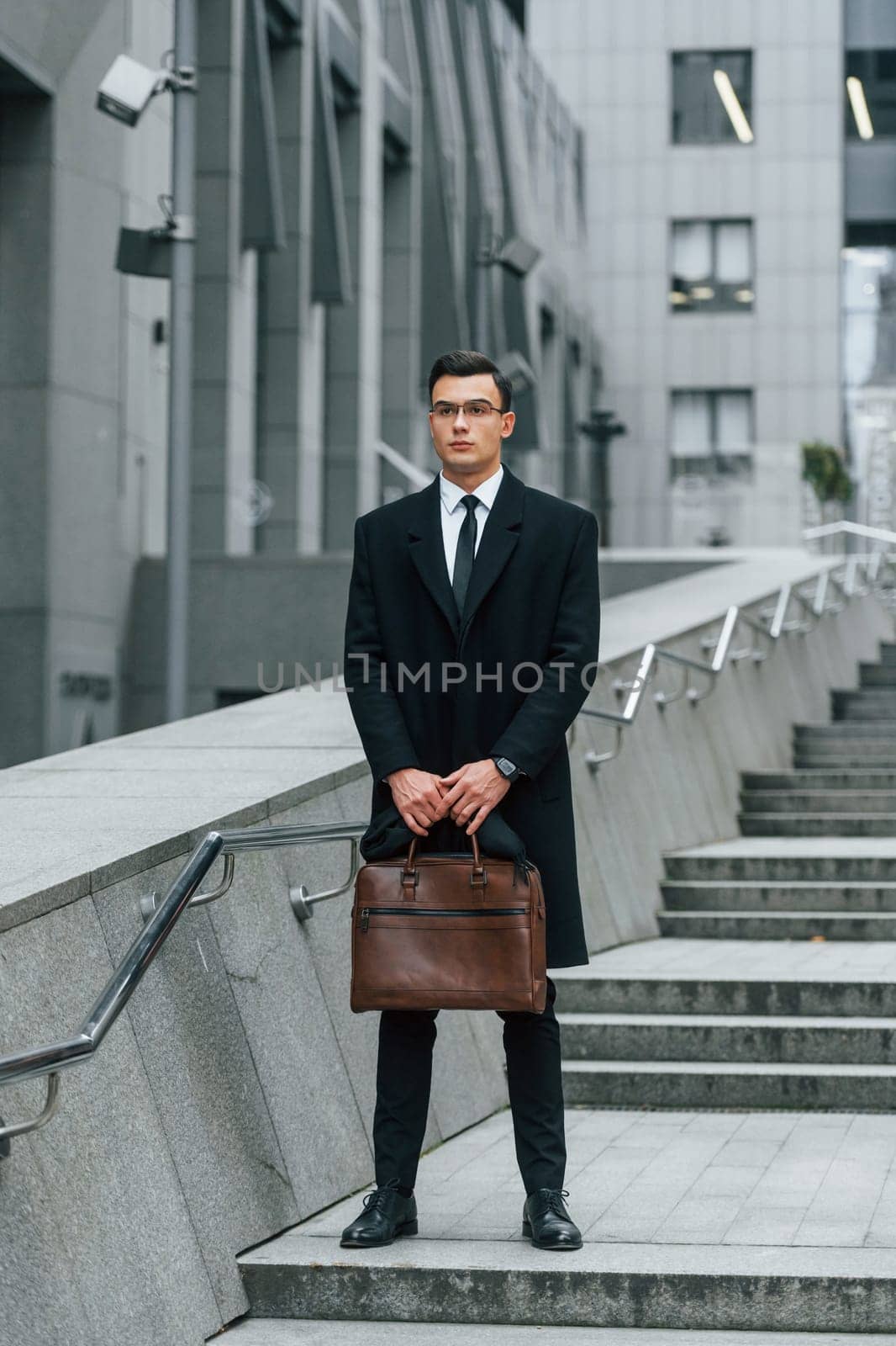 Holding brown bag in hands. Businessman in black suit and tie is outdoors in the city.