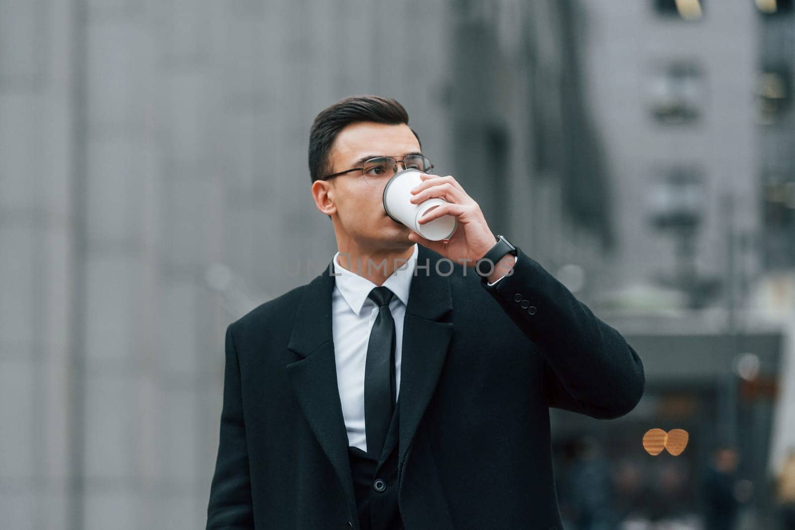 With cup of drink. Businessman in black suit and tie is outdoors in the city by Standret