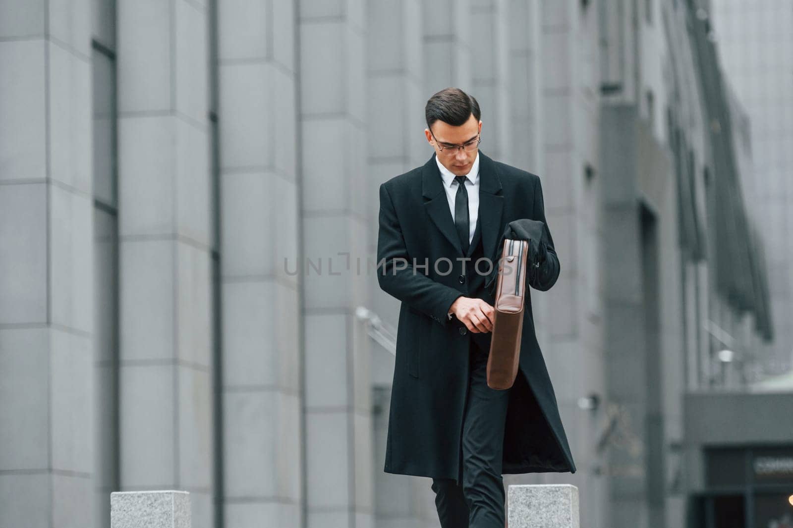 Holding brown bag in hands. Businessman in black suit and tie is outdoors in the city.
