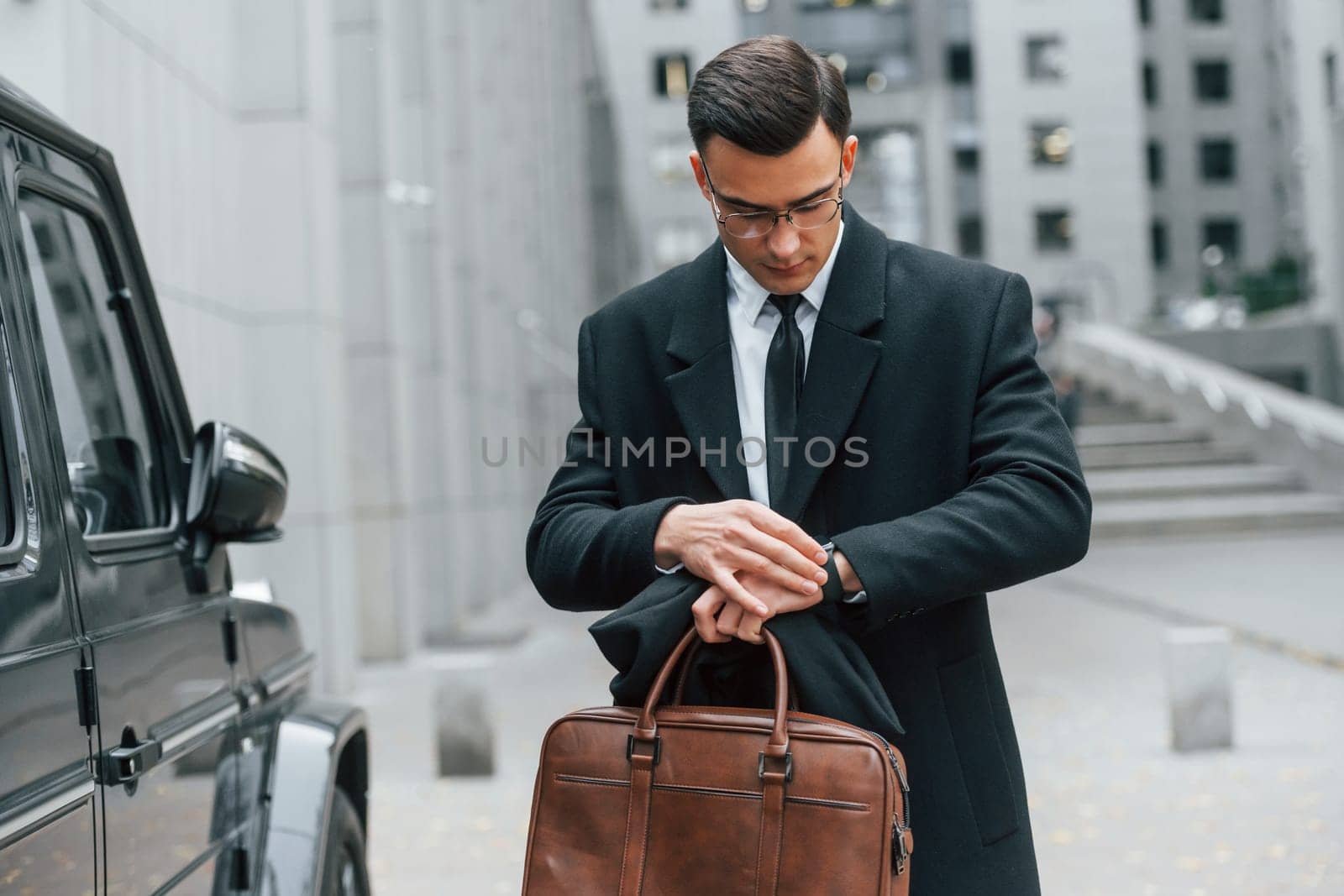 Holding brown bag in hands. Businessman in black suit and tie is outdoors in the city.