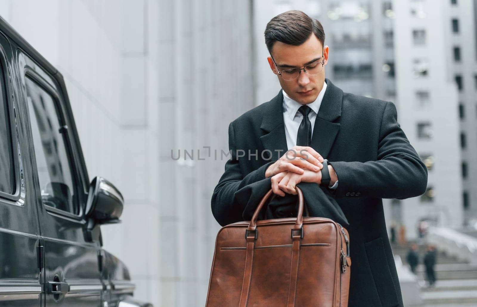 Holding brown bag in hands. Businessman in black suit and tie is outdoors in the city.