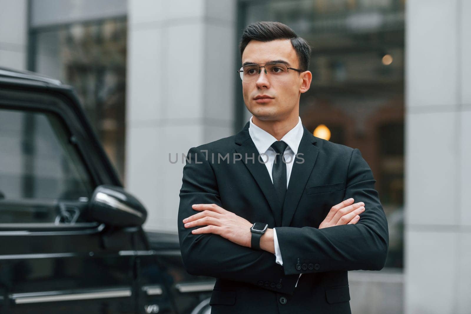 Standing with arms crossed. Businessman in black suit and tie is outdoors in the city.