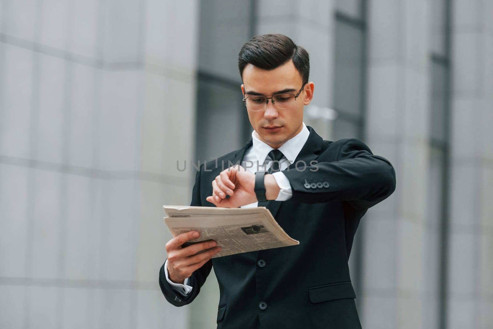 Holding newspaper. Businessman in black suit and tie is outdoors in the city.