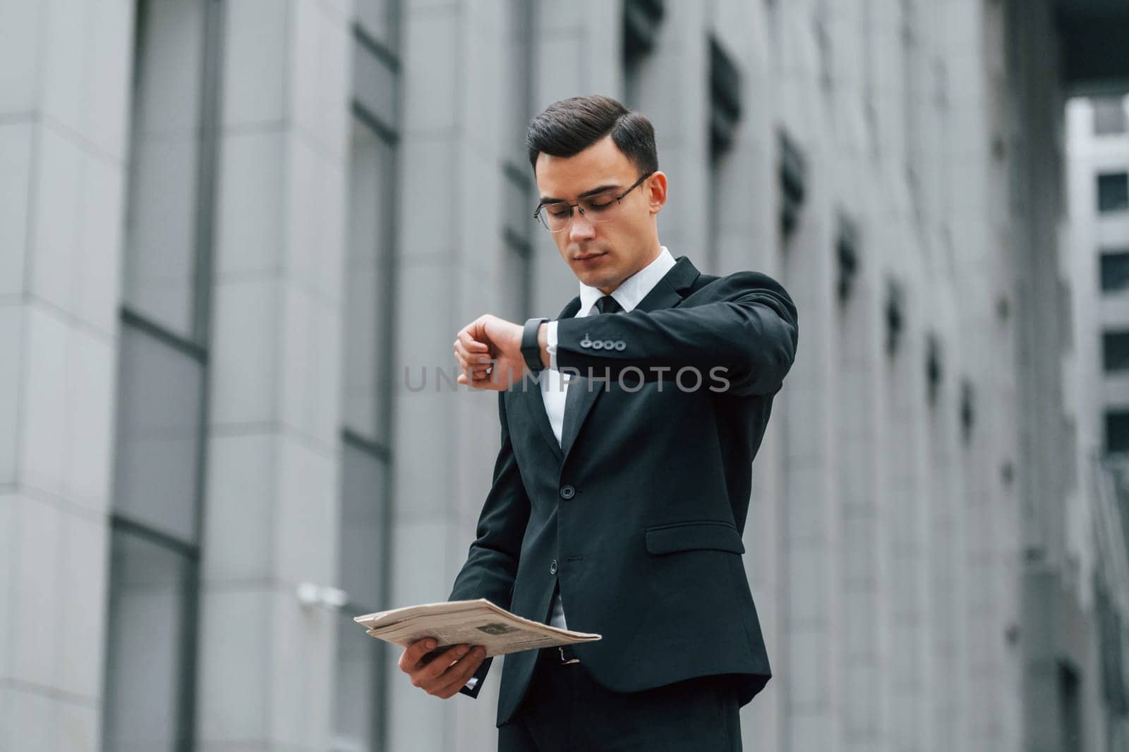 Holding newspaper. Businessman in black suit and tie is outdoors in the city by Standret