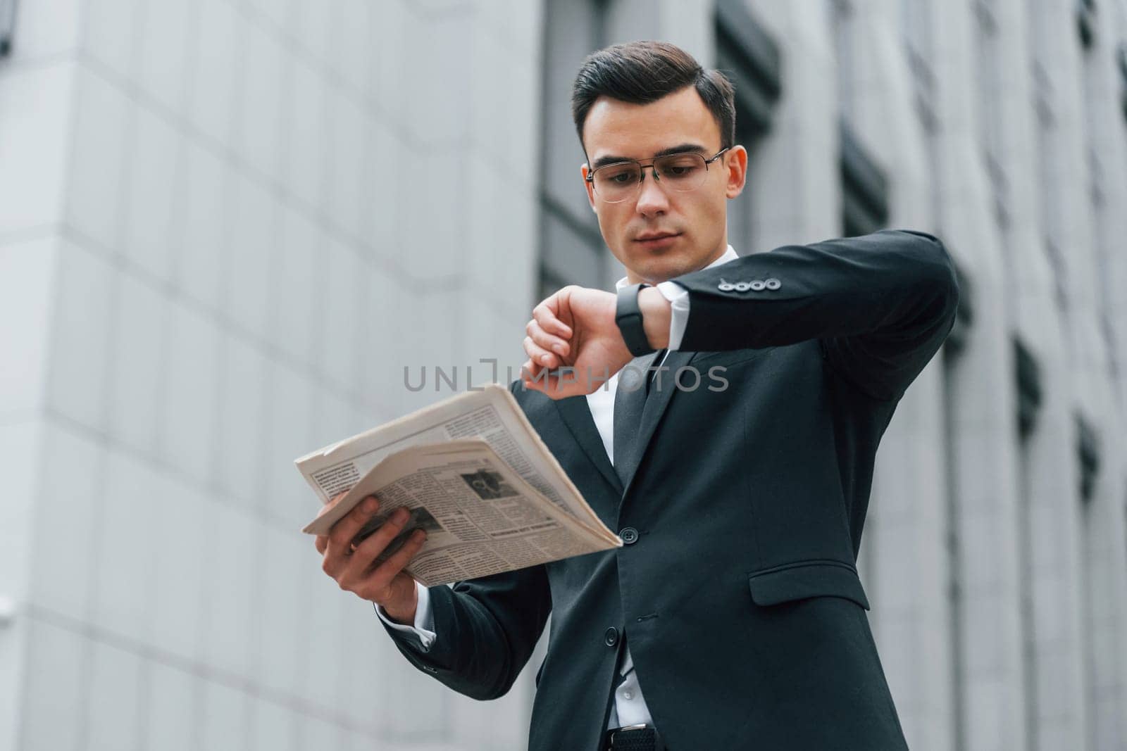 Holding newspaper. Businessman in black suit and tie is outdoors in the city.