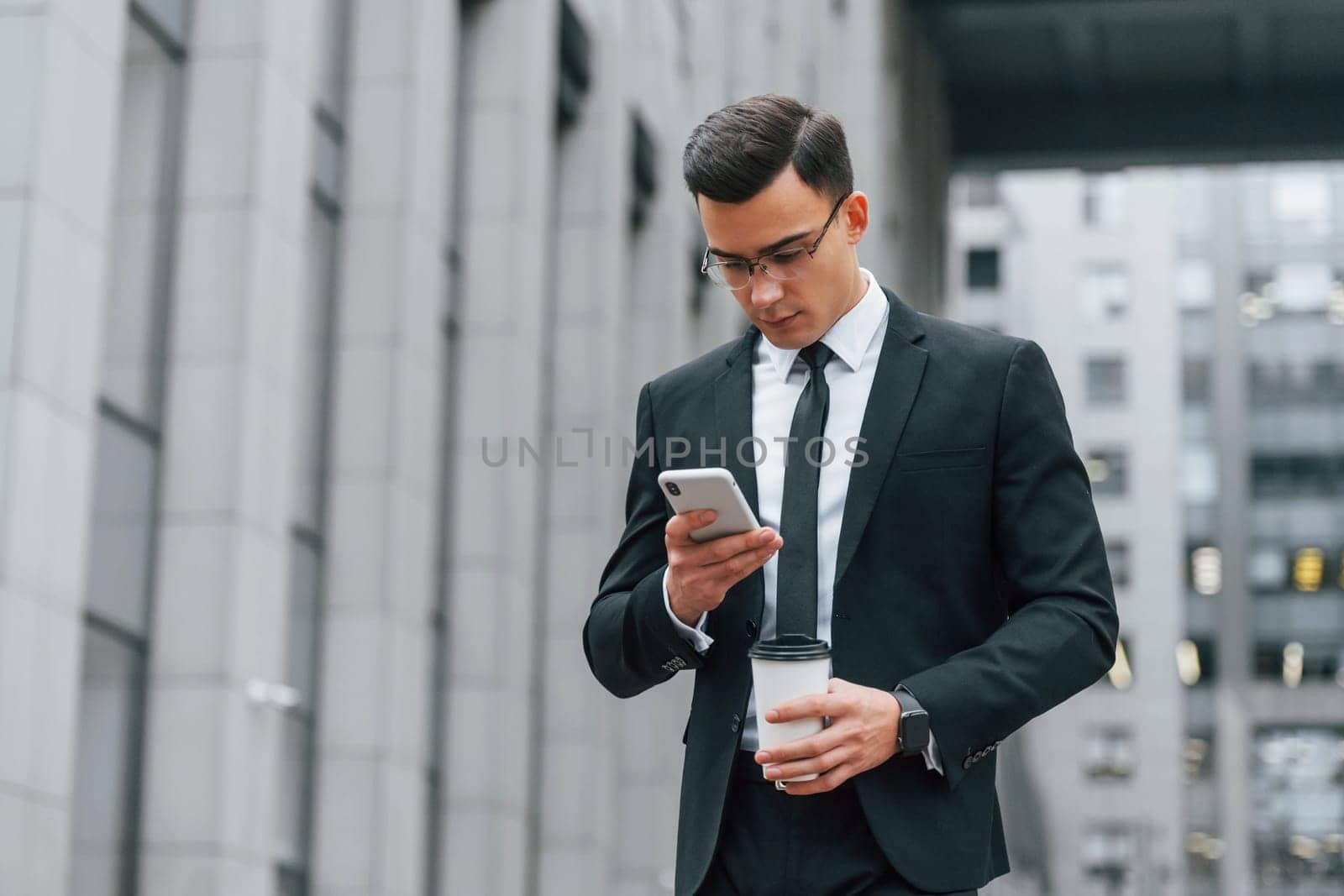 Using phone. Businessman in black suit and tie is outdoors in the city.