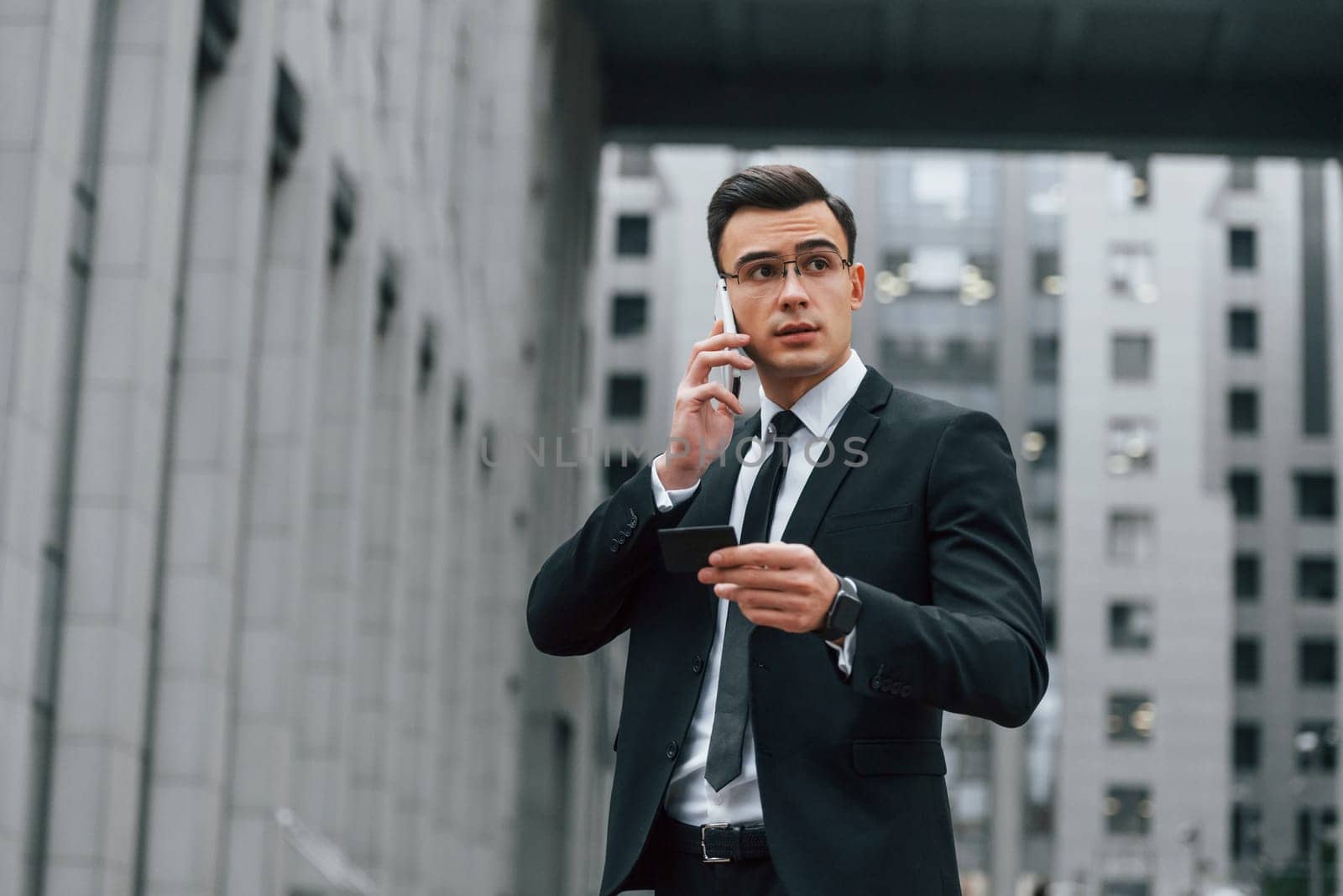 Using phone. Businessman in black suit and tie is outdoors in the city by Standret