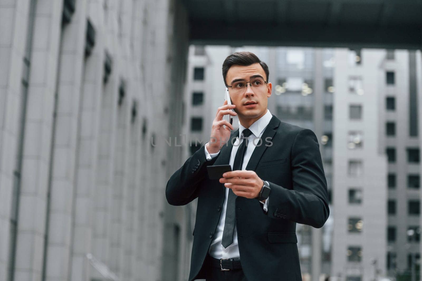 Using phone. Businessman in black suit and tie is outdoors in the city.