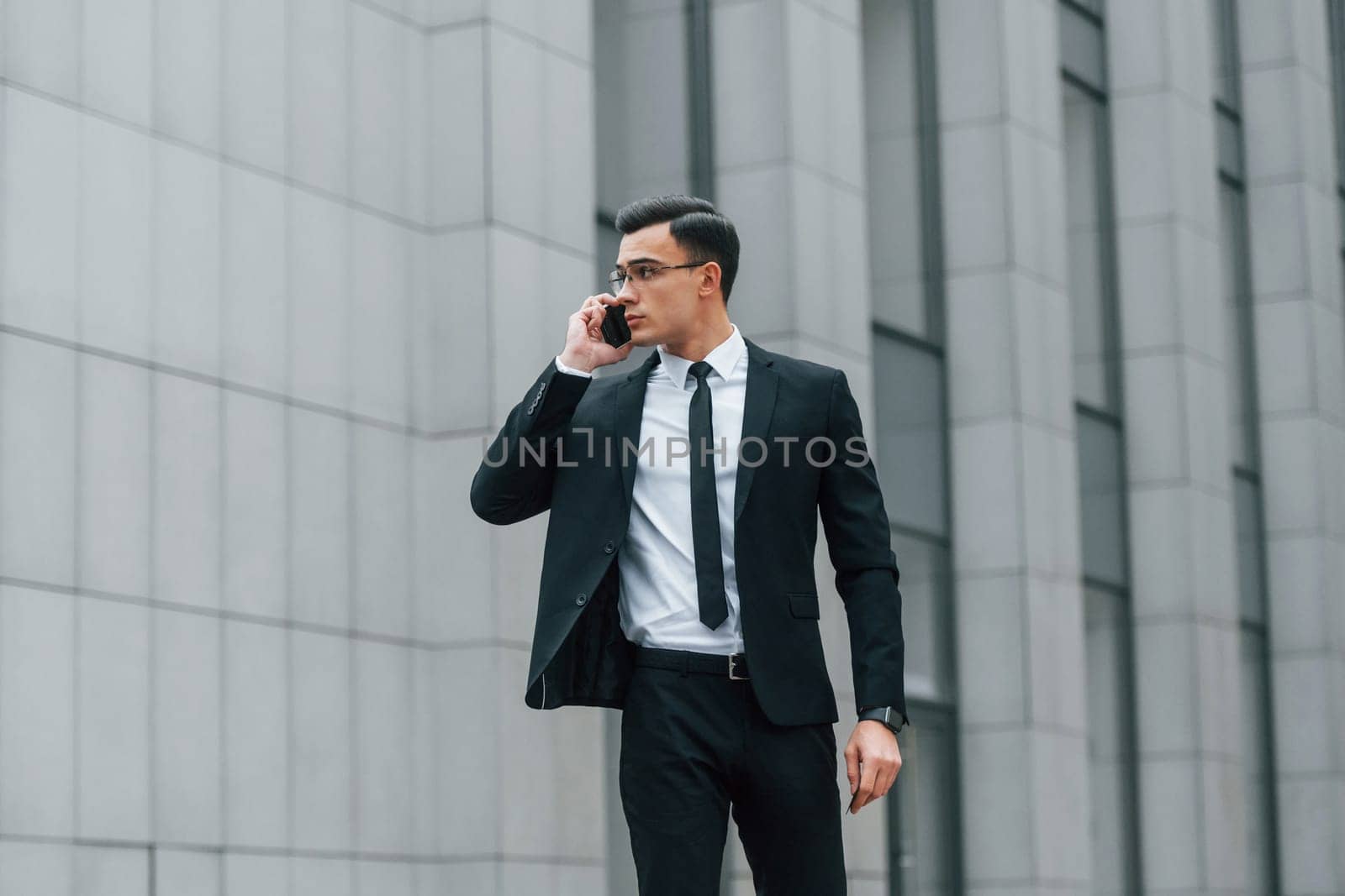 Using phone. Businessman in black suit and tie is outdoors in the city.