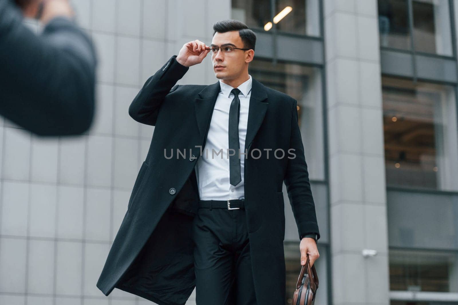 Posing for photographer. Businessman in black suit and tie is outdoors in the city.