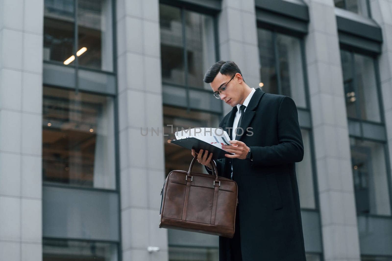 Holding documents. Businessman in black suit and tie is outdoors in the city by Standret