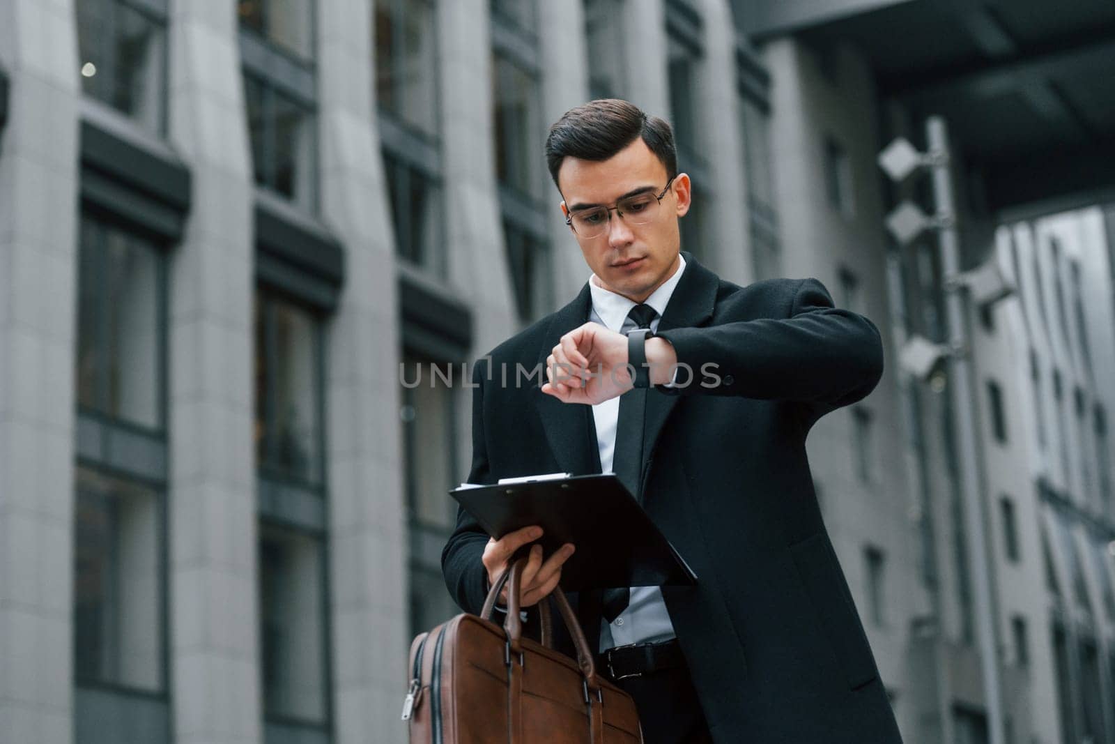 Looking at clock. Businessman in black suit and tie is outdoors in the city.