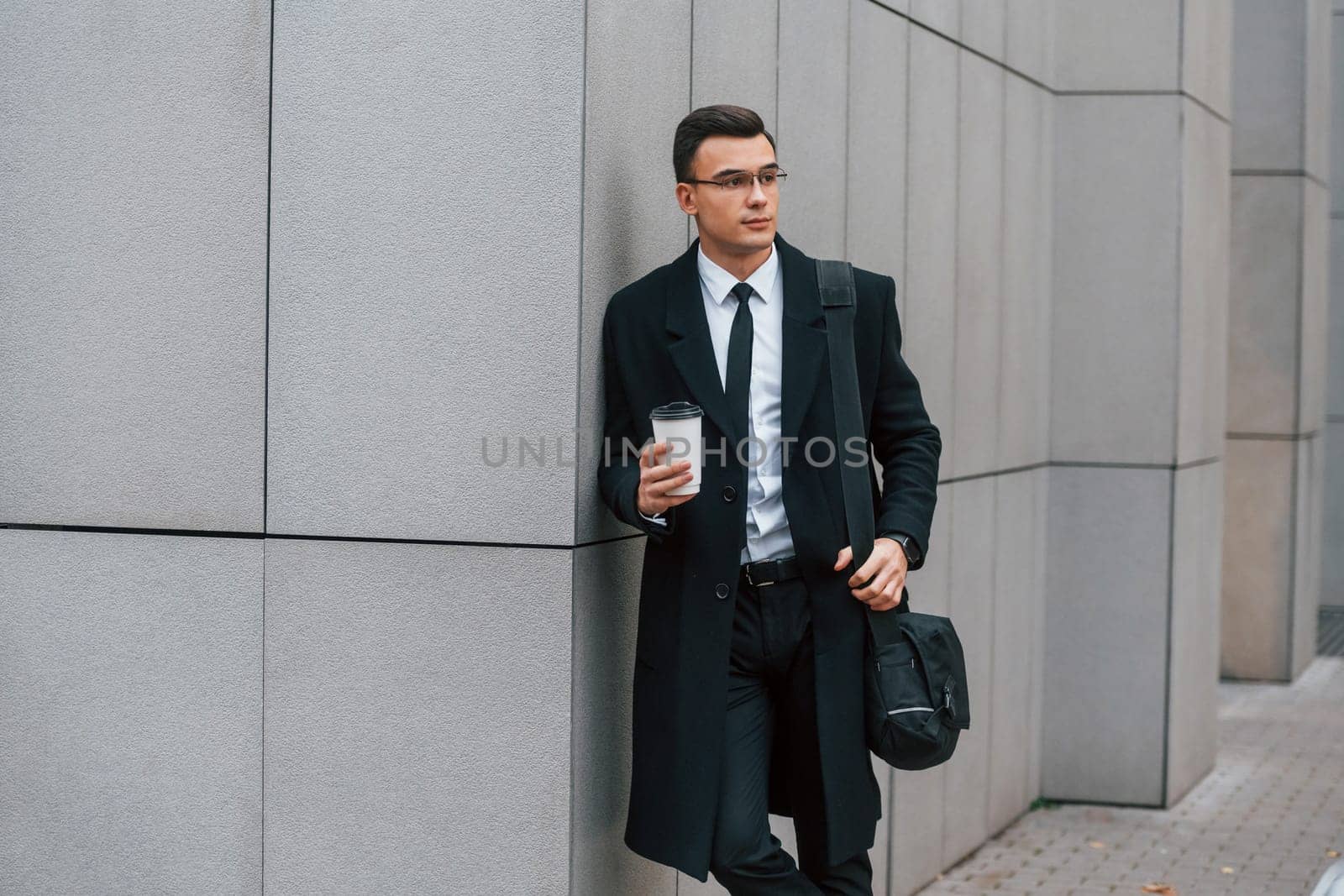 With cup of drink. Businessman in black suit and tie is outdoors in the city.