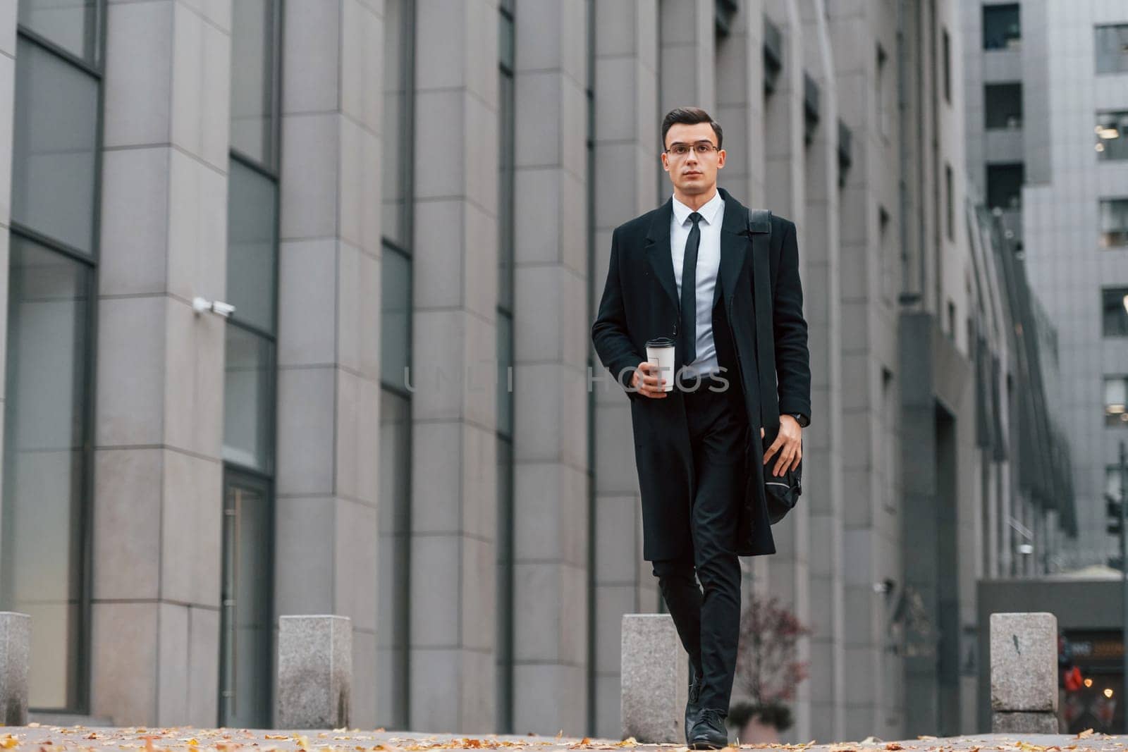 With cup of drink. Businessman in black suit and tie is outdoors in the city.