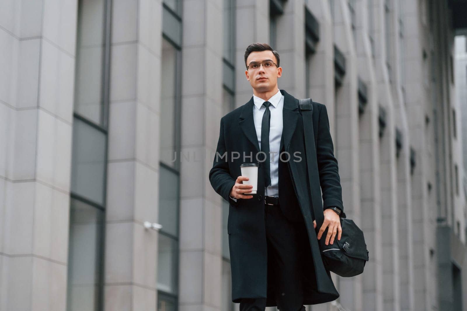 With cup of drink. Businessman in black suit and tie is outdoors in the city.